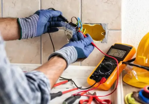 A close-up of an electrician wearing gloves, working with exposed wires and a socket inside a wall. A digital multimeter displays a voltage reading, with various tools and a yellow hard hat nearby.